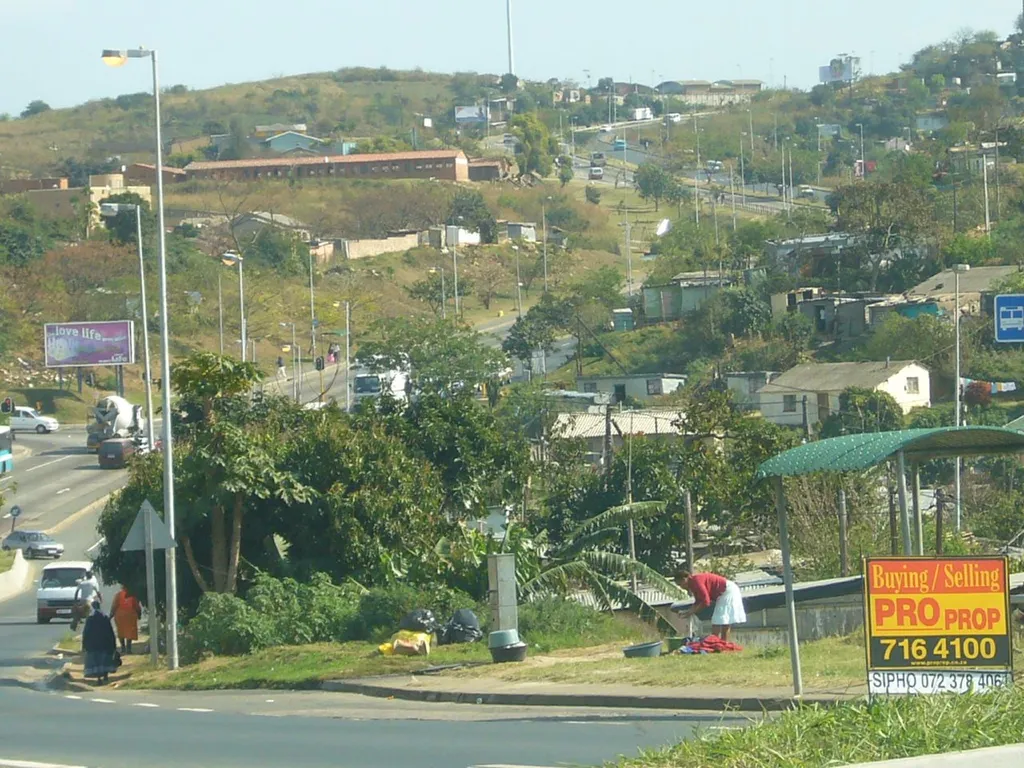 A street in Umlazi township near Durban, KwaZulu-Natal, South Africa, with houses on a hillside and a woman washing laundry at a public tap.