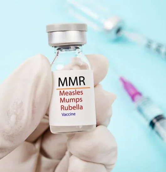 A gloved hand holds a vial labelled MMR — Measles Mumps Rubella vaccine, with a syringe in the background.