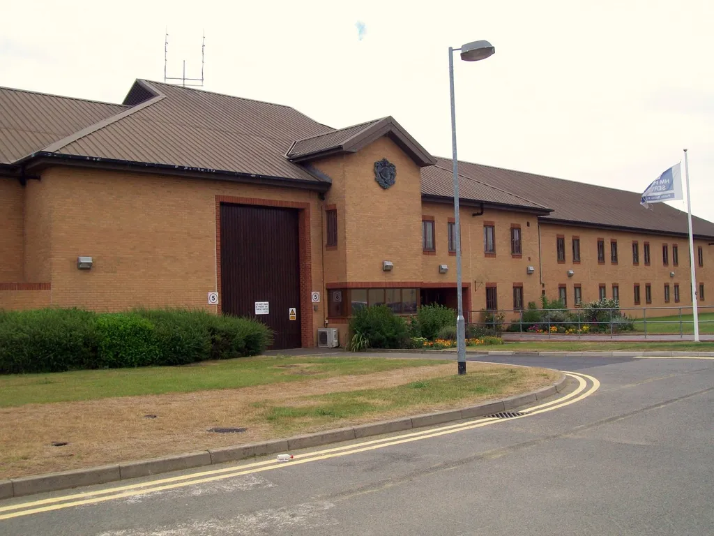 The brick exterior of HMP Littlehey prison in Cambridgeshire, showing the main entrance gate, the Prison Service crest and an HM Prison Service flag flying from a flagpole.