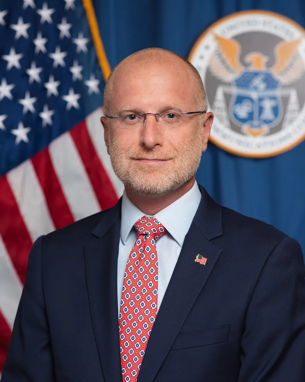 Official portrait of FCC Chairman Brendan Carr in a navy suit and red tie against a dark background