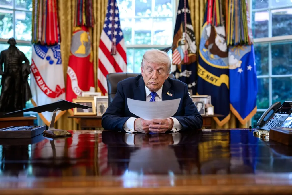 President Donald Trump seated at the Resolute desk in the Oval Office reading a document, with the US flag and military service flags behind him