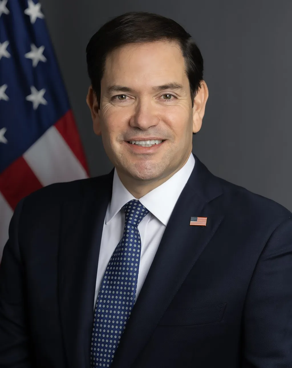 Official portrait of US Secretary of State Marco Rubio in a dark suit and blue tie, with a US flag pin on his lapel and the American flag behind him
