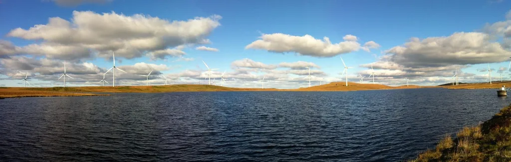 Dozens of white wind turbines with turning blades spread across moorland hills around a reservoir under a blue sky with scattered clouds