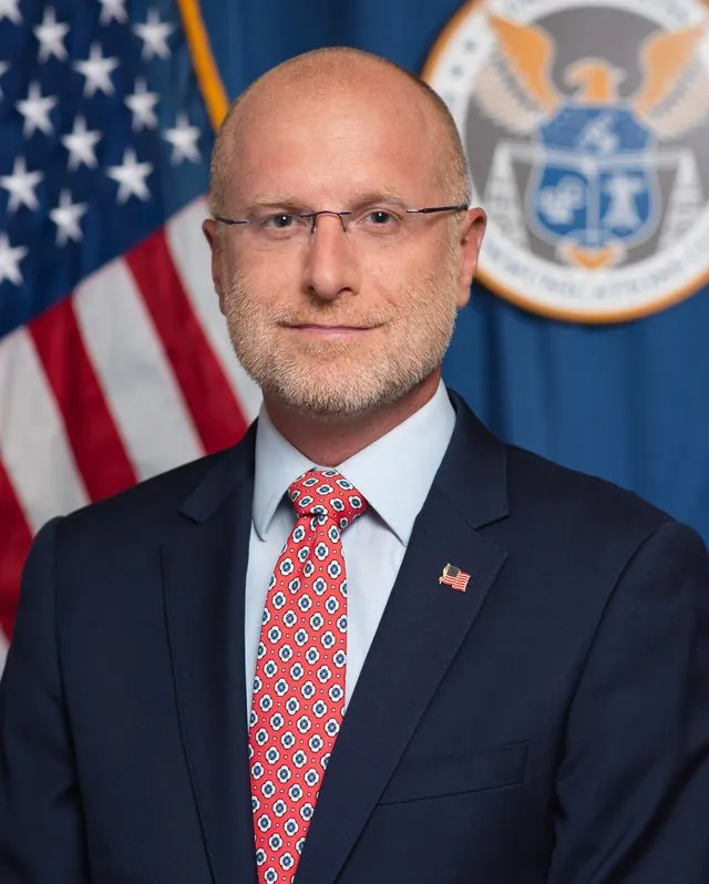 Official portrait of FCC Chair Brendan Carr, an adult man with short brown hair in a dark suit and blue tie against a dark background.