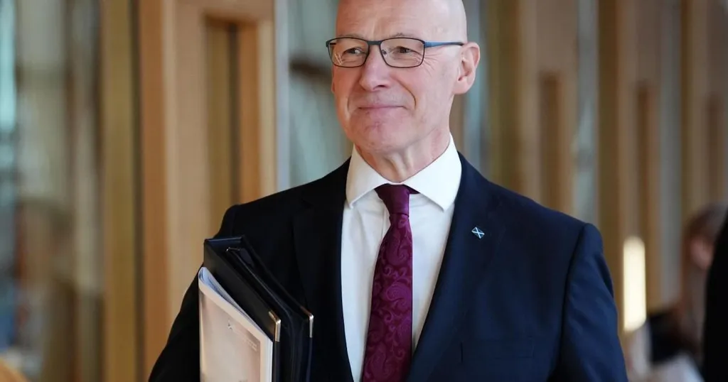 John Swinney holding folders and smiling, wearing a suit with a Scotland Saltire pin