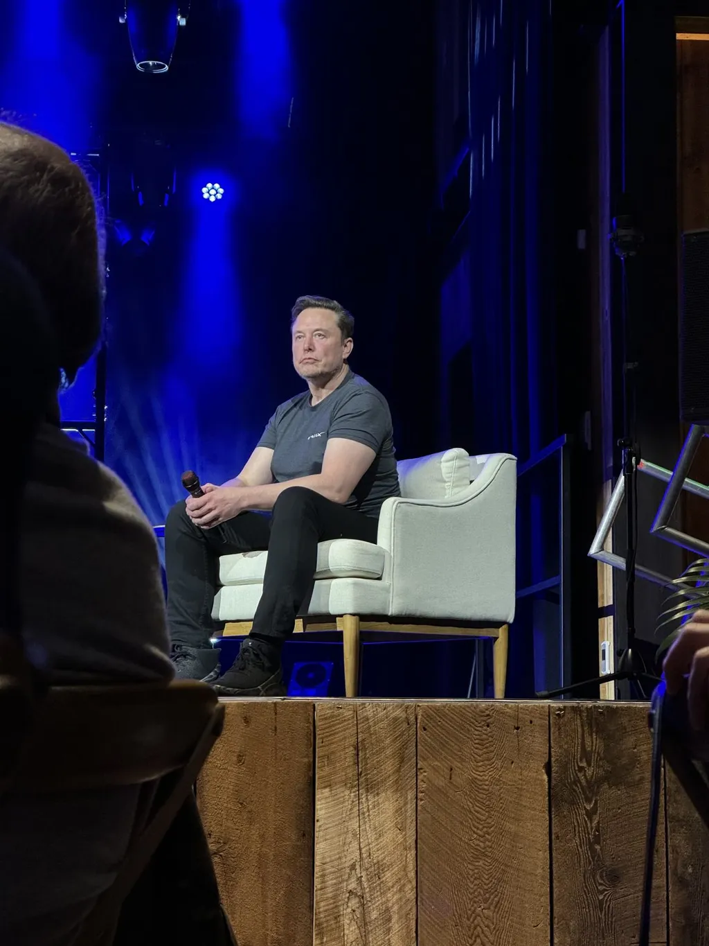 Elon Musk seated on a stage chair holding a microphone at a conference in March 2024