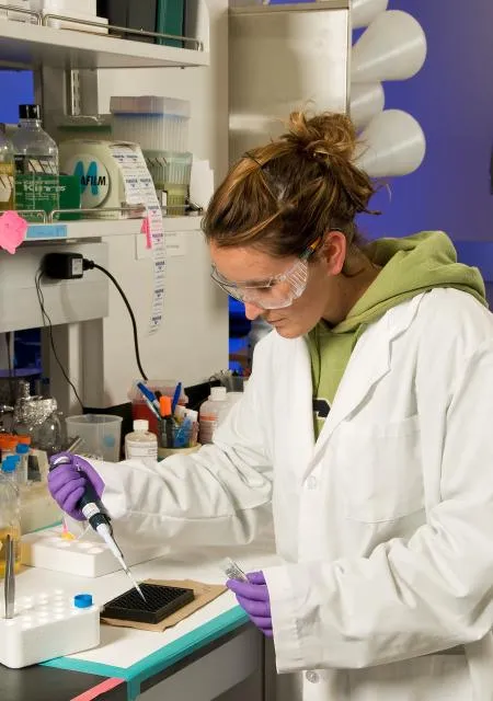 A laboratory researcher in a white coat and safety glasses pipetting samples into a microplate
