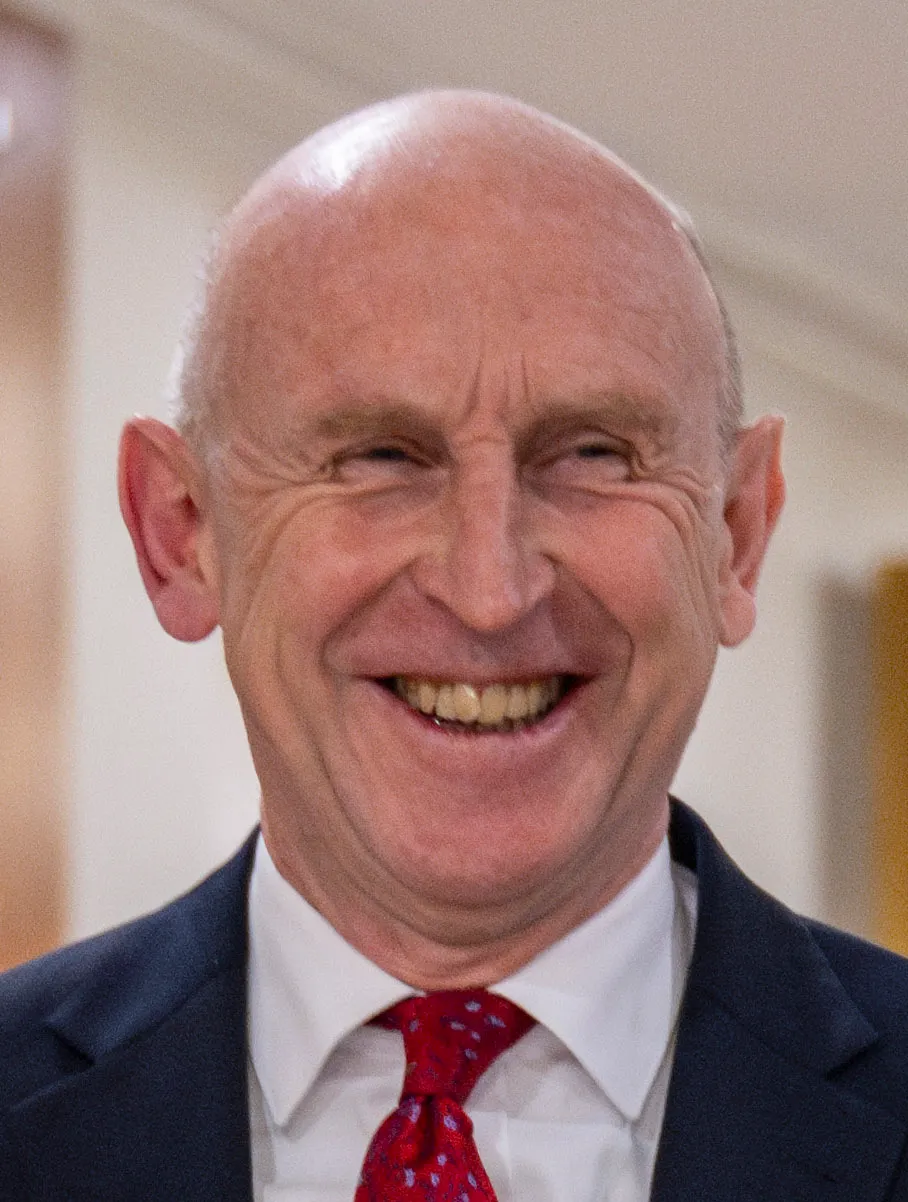 Portrait of Chancellor John Healey in a dark suit and red tie, smiling.