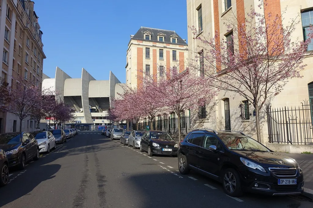 The Parc des Princes stadium in Paris, its distinctive concrete architecture visible at the end of a quiet street lined with flowering trees