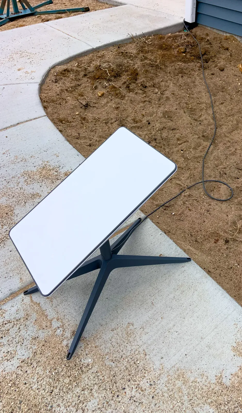 A white rectangular Starlink satellite dish on a black tripod stand with cable, on outdoor ground