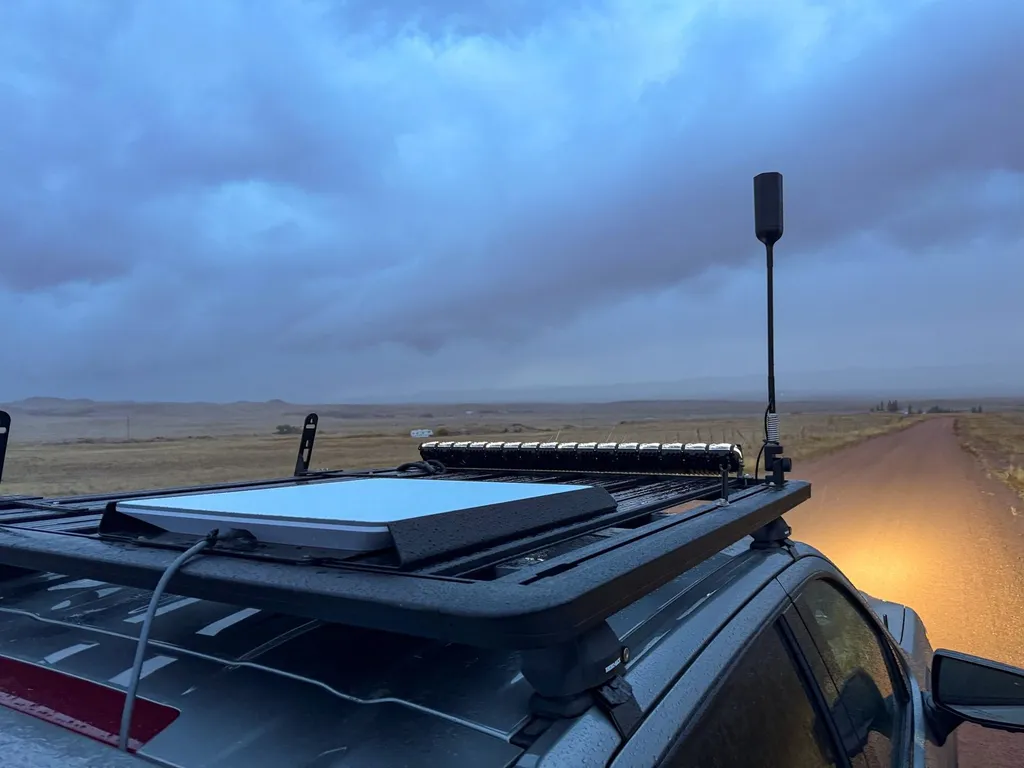 A Starlink flat-panel satellite dish mounted on the roof rack of an overland truck on a rural dirt road under a cloudy evening sky.
