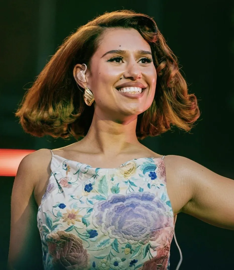 RAYE smiling on stage in a floral dress, performing in 2025