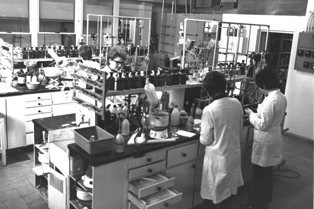 Scientists in white coats at work inside a pharmaceutical research laboratory, with benches of glassware and chemical bottles (archival photograph)