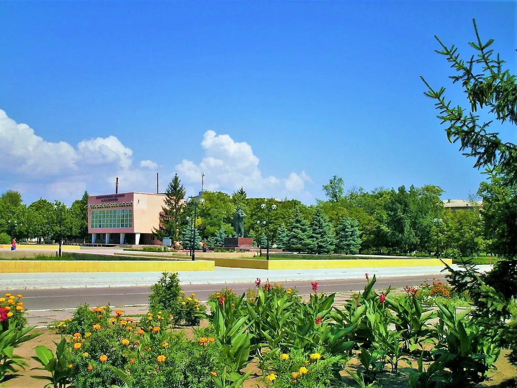 Central square of Oleshky in Kherson Oblast — flower beds, a statue and a civic building under a blue sky