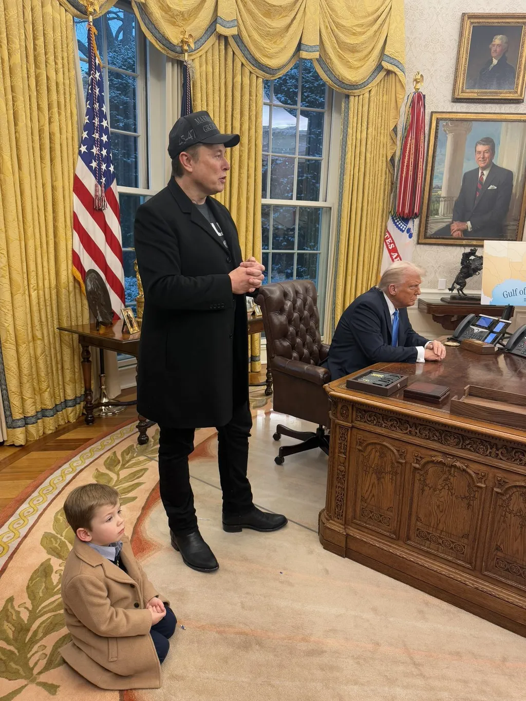 Elon Musk standing in the Oval Office beside the Resolute desk where President Donald Trump is seated, both taking questions from the press.