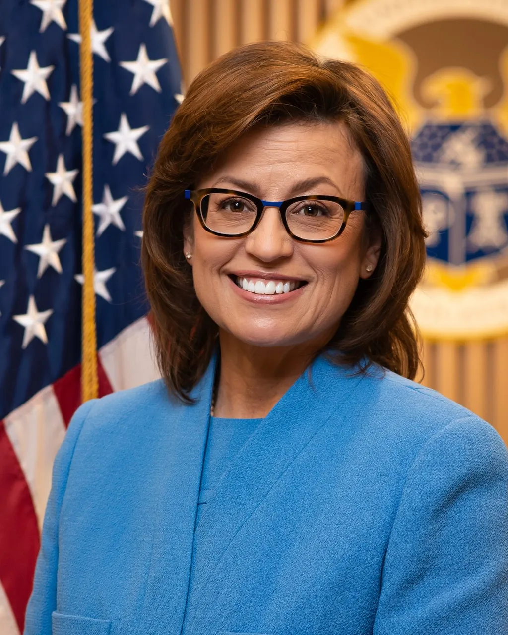 Official FCC portrait of Commissioner Anna Gomez, wearing glasses and a blue blazer, in front of the US flag and the FCC seal