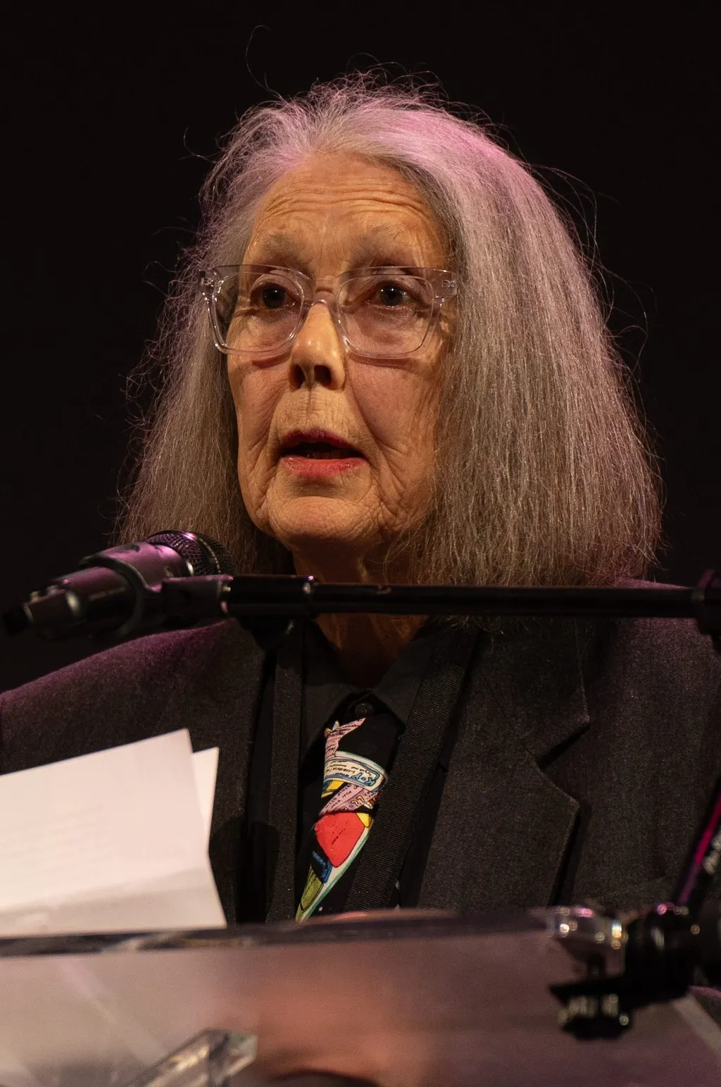 Portrait of Canadian poet Anne Carson, an elderly woman with long grey hair and glasses, reading from papers at a microphone at the 2024 National Book Awards finalist reading.