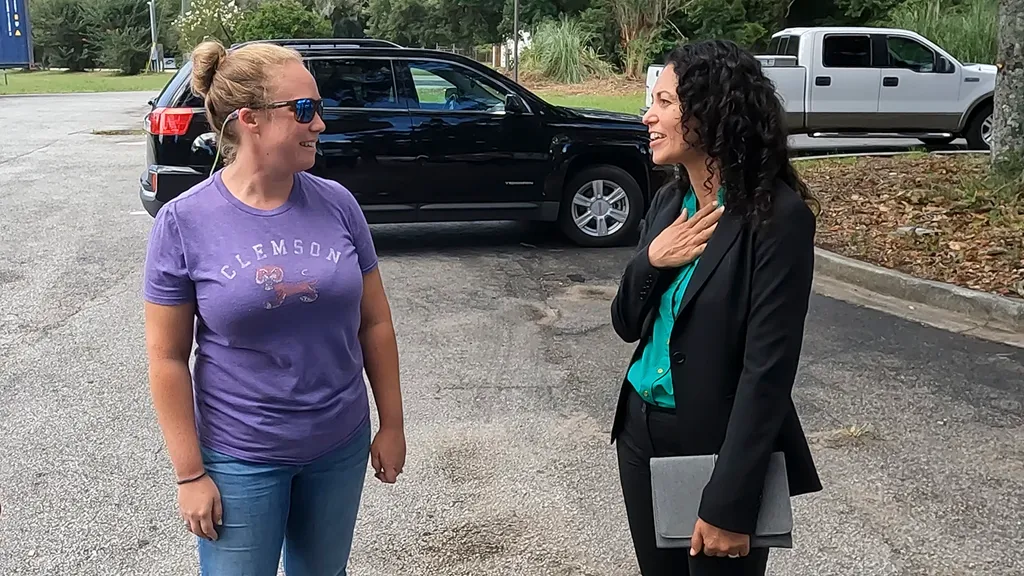 Two women talking in a car park — one in a purple Clemson T-shirt and jeans, the other in a black blazer over a teal shirt, holding a folder.