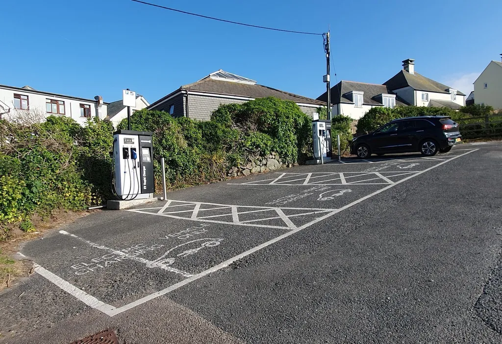 An EV rapid charging point beside a marked 'electric vehicle charging only' bay in a UK car park, with houses and a black car nearby
