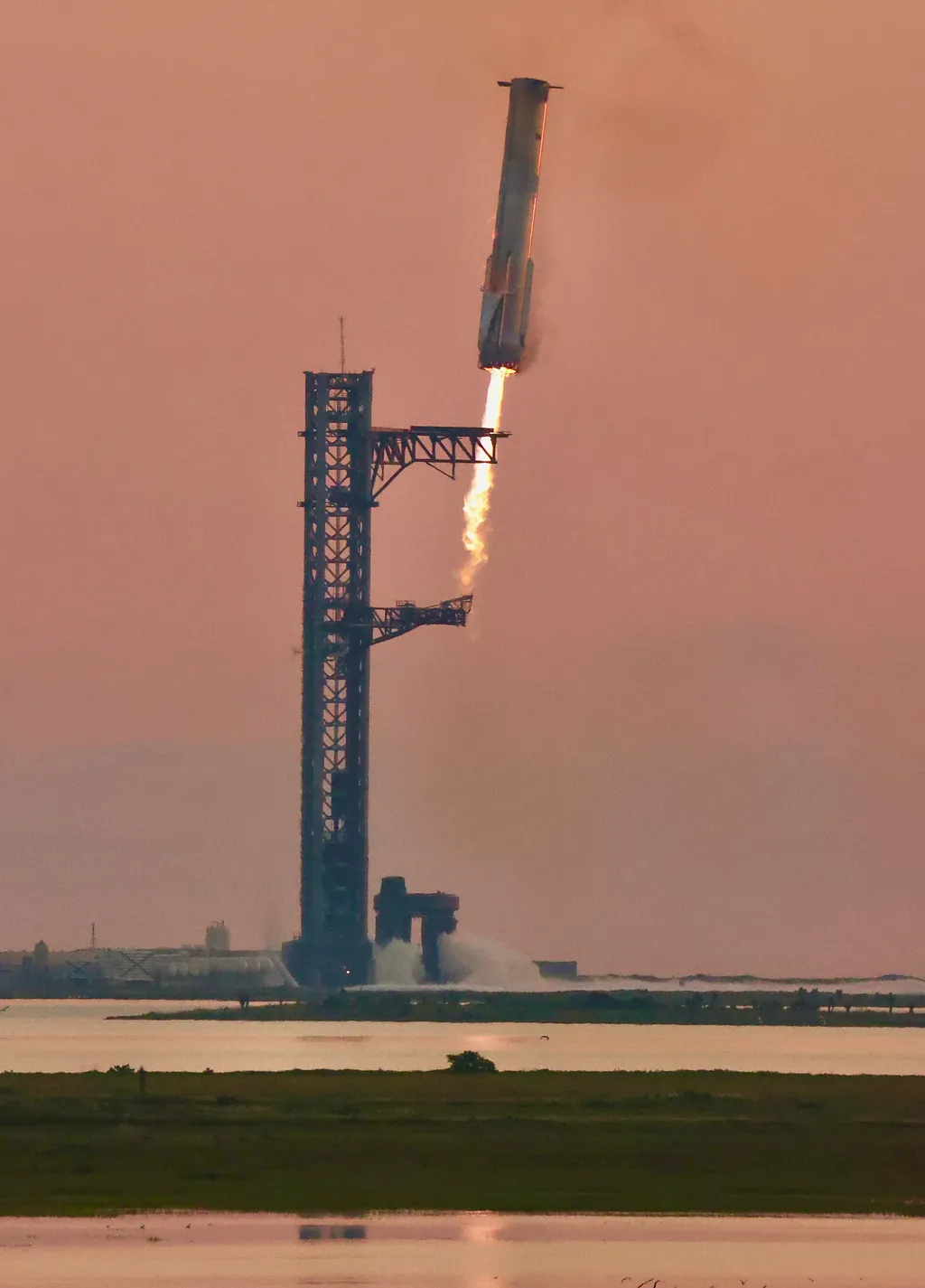 A SpaceX Starship booster descending to the launch tower for a chopsticks catch at sunset, engines flaring.