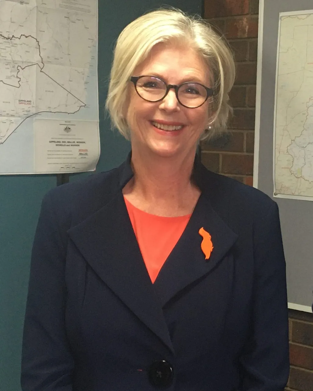 Portrait of Australian independent MP Helen Haines, smiling, wearing glasses and a navy blazer with an orange lapel pin, in front of electorate maps