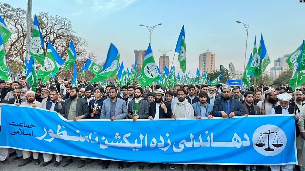 Jamaat-e-Islami supporters marching in Islamabad, carrying green-and-blue party flags bearing the scales-of-justice emblem and a large blue banner with Urdu text rejecting the 2024 election results (file photo from 2024).