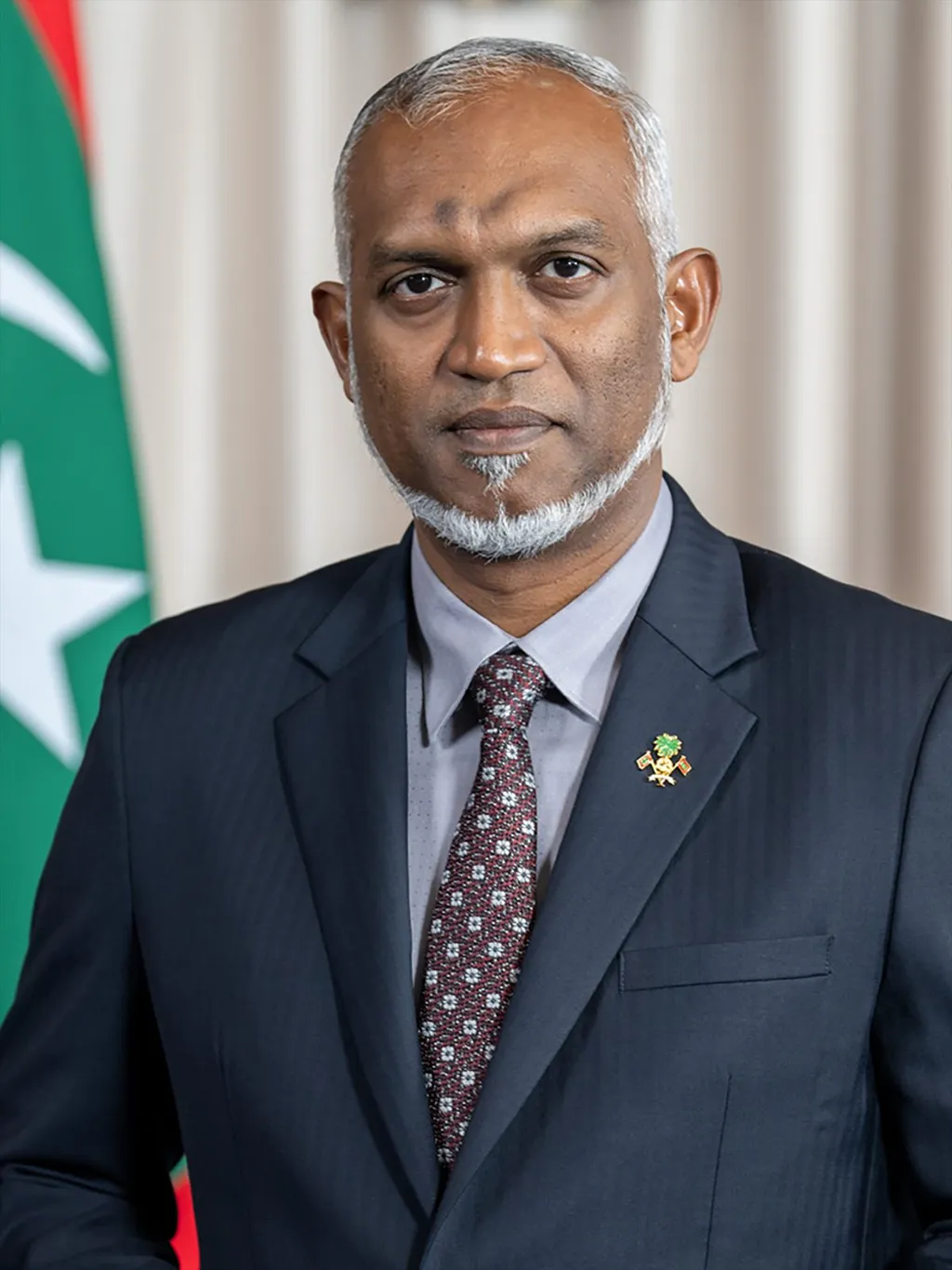 Official portrait of Maldivian President Dr Mohamed Muizzu, wearing a dark suit with a Maldivian national emblem lapel pin, the Maldives flag behind him.
