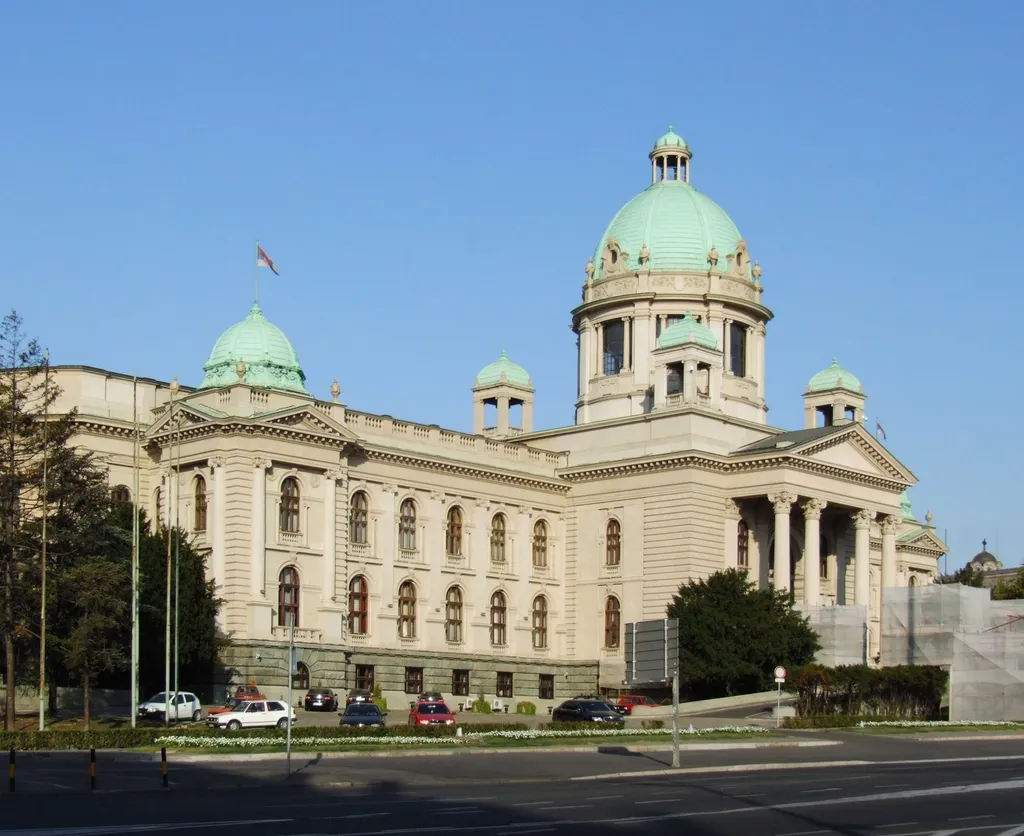 The National Assembly of Serbia building in Belgrade, a neoclassical stone building with a green copper dome and colonnaded portico, photographed in daylight under a clear blue sky.