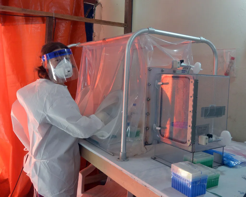 An NIAID researcher in full protective equipment works at a sealed glovebox in an Ebola diagnostic laboratory, Monrovia, handling patient blood samples.