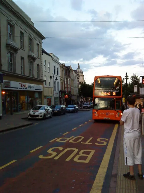 A red Nottingham city bus stopped at a bus stop on Angel Row, Nottingham city centre, with a Tesco Express storefront visible on the street (archive photo of the location)