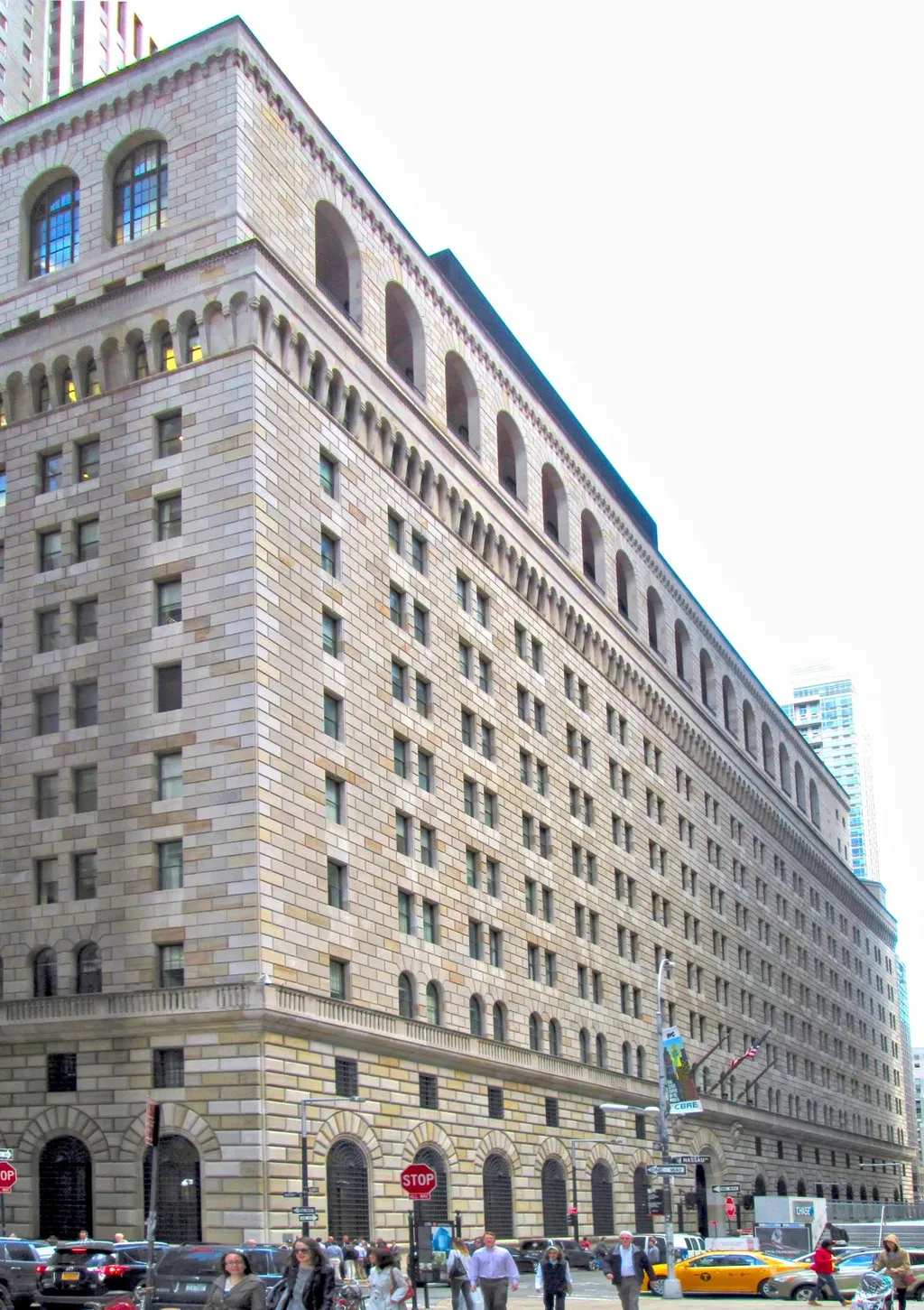 The Federal Reserve Bank of New York building at 33 Liberty Street in Manhattan, viewed from the west, showing its rusticated limestone facade with the double-height arched main entrance and wrought-iron lanterns.