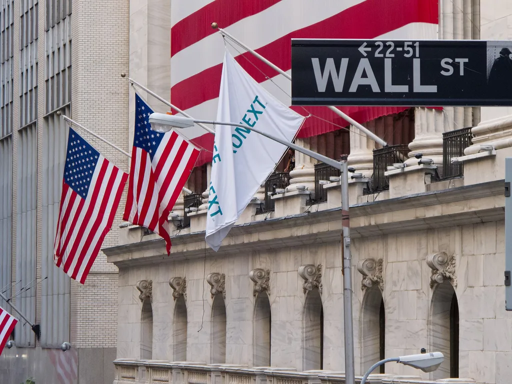 The facade of the New York Stock Exchange building on Wall Street, with US flags and the Wall Street sign visible.
