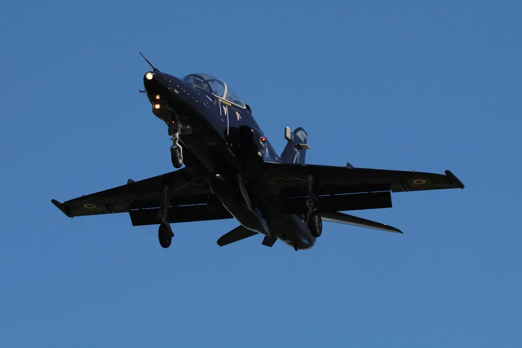 An RAF Hawk T2 training jet at RAF Valley in flight, with landing gear down against a clear blue sky.