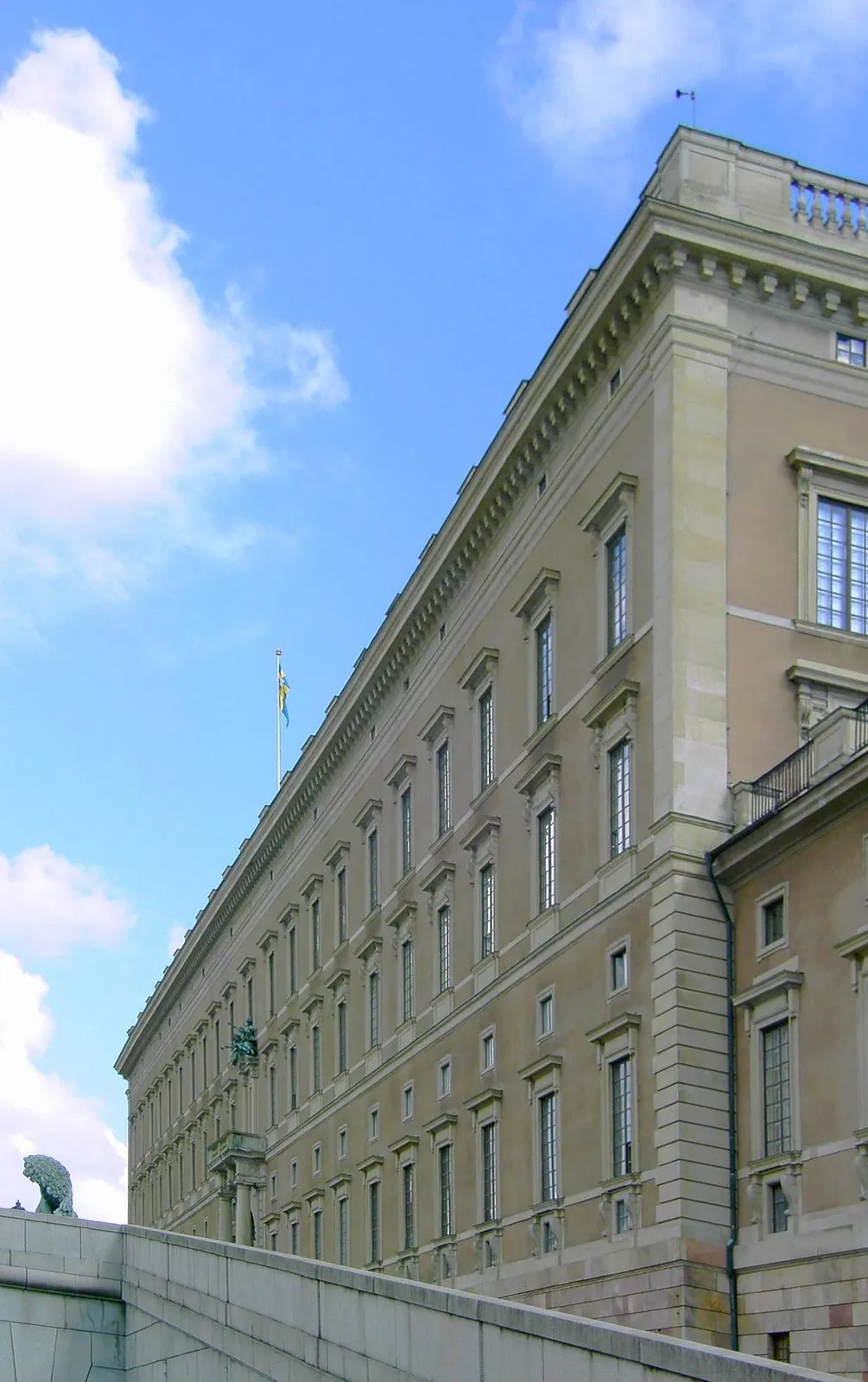 The sandstone baroque facade of the Royal Palace in Stockholm photographed from Lejonbacken, with a Swedish flag flying above it and a stone lion statue in the foreground