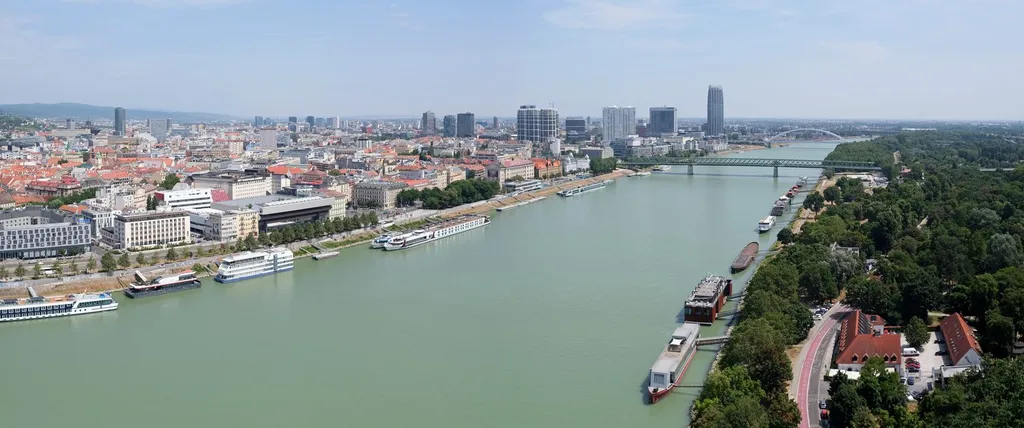 The Danube riverfront at Bratislava, Slovakia: the Old Bridge spans the river with the old-town rooftops and modern city skyline behind, moored river cruise boats in the foreground.