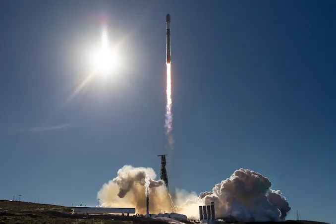 A Falcon 9 rocket lifting off in daylight from Vandenberg, trailing a bright plume of fire and smoke.