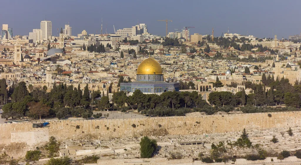View of the Old City of Jerusalem from the Mount of Olives, with the golden dome of the Dome of the Rock rising above the city walls and rooftops under a clear sky.