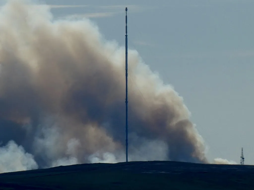 Smoke from a wildfire rises over moorland on Winter Hill, Lancashire, with the Winter Hill transmitting mast in the foreground.