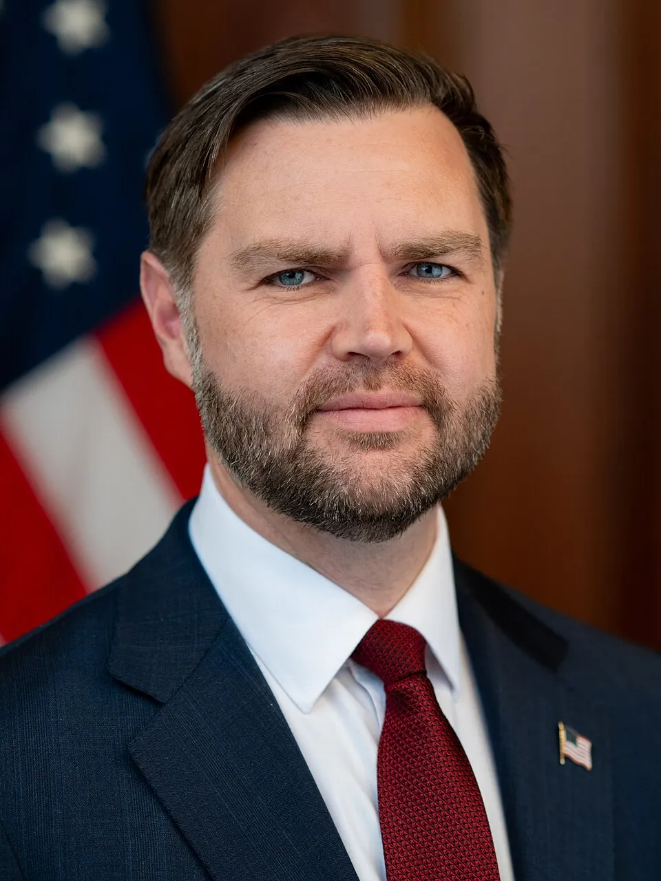 Official March 2026 portrait of US Vice President JD Vance in a dark suit, white shirt and red tie with an American flag pin, standing before the American flag