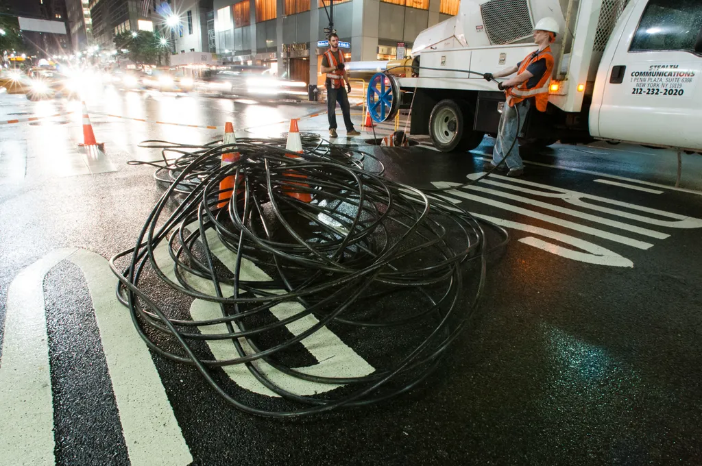 Fibre-optic cable installation on a city street at night: workers feed a coiled cable from a service van amid traffic cones.